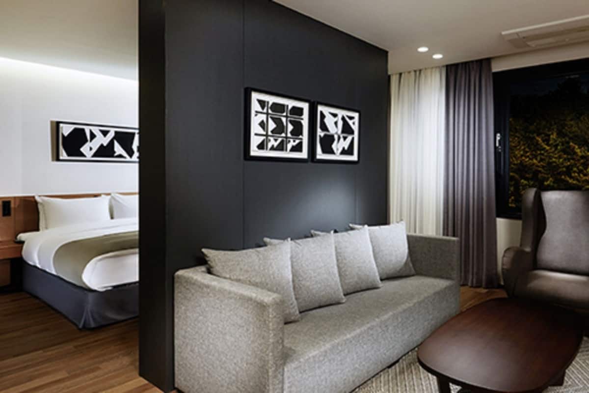 A modern hotel room with a bed, a grey sofa, a brown armchair, and a desk. There are abstract wall art pieces and a window with curtains.