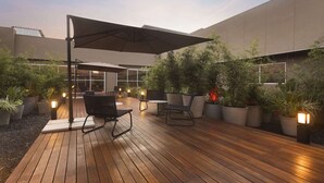 Un patio al aire libre con suelo de madera, mobiliario, plantas en macetas y un gran paraguas que proporciona sombra.