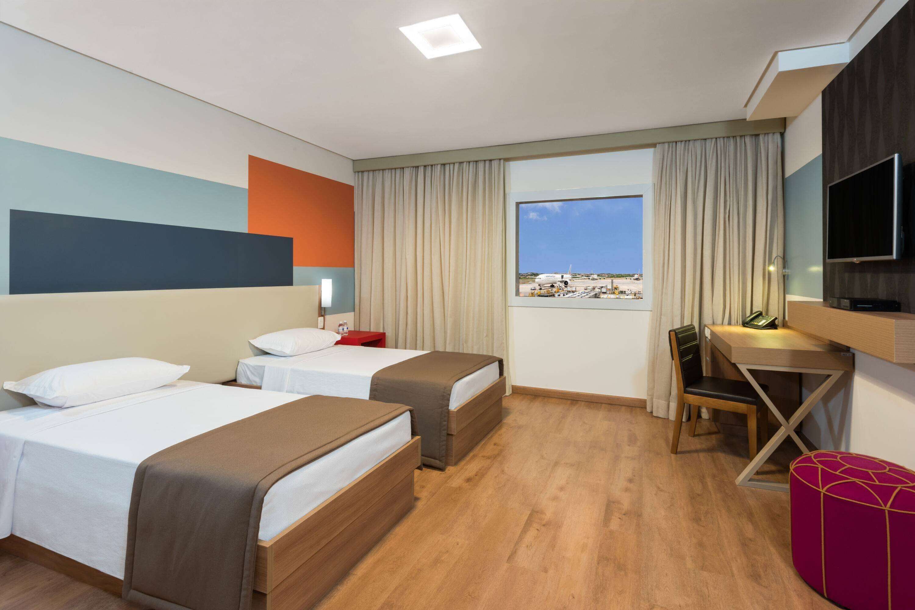 A hotel room with two beds, a desk, a chair, a TV, and a window with a beach view.