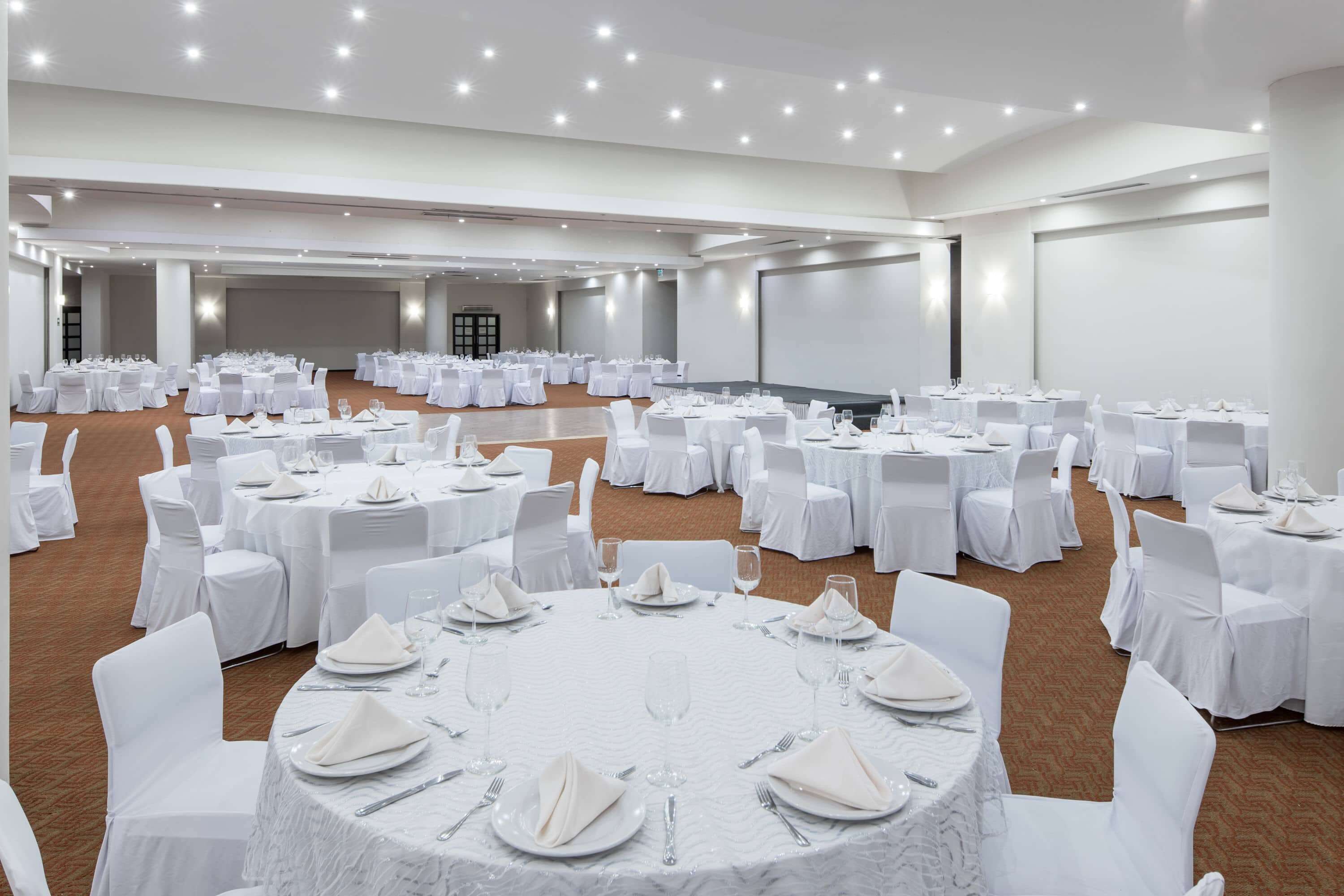 Un amplio salón de conferencias con mesas redondas dispuestas para comedor, sillas blancas y suelo alfombrado.
