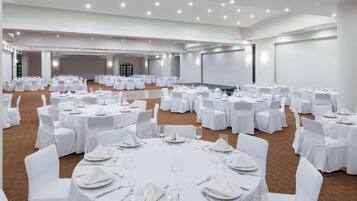 Un amplio salón de conferencias con mesas redondas dispuestas para comedor, sillas blancas y suelo alfombrado.