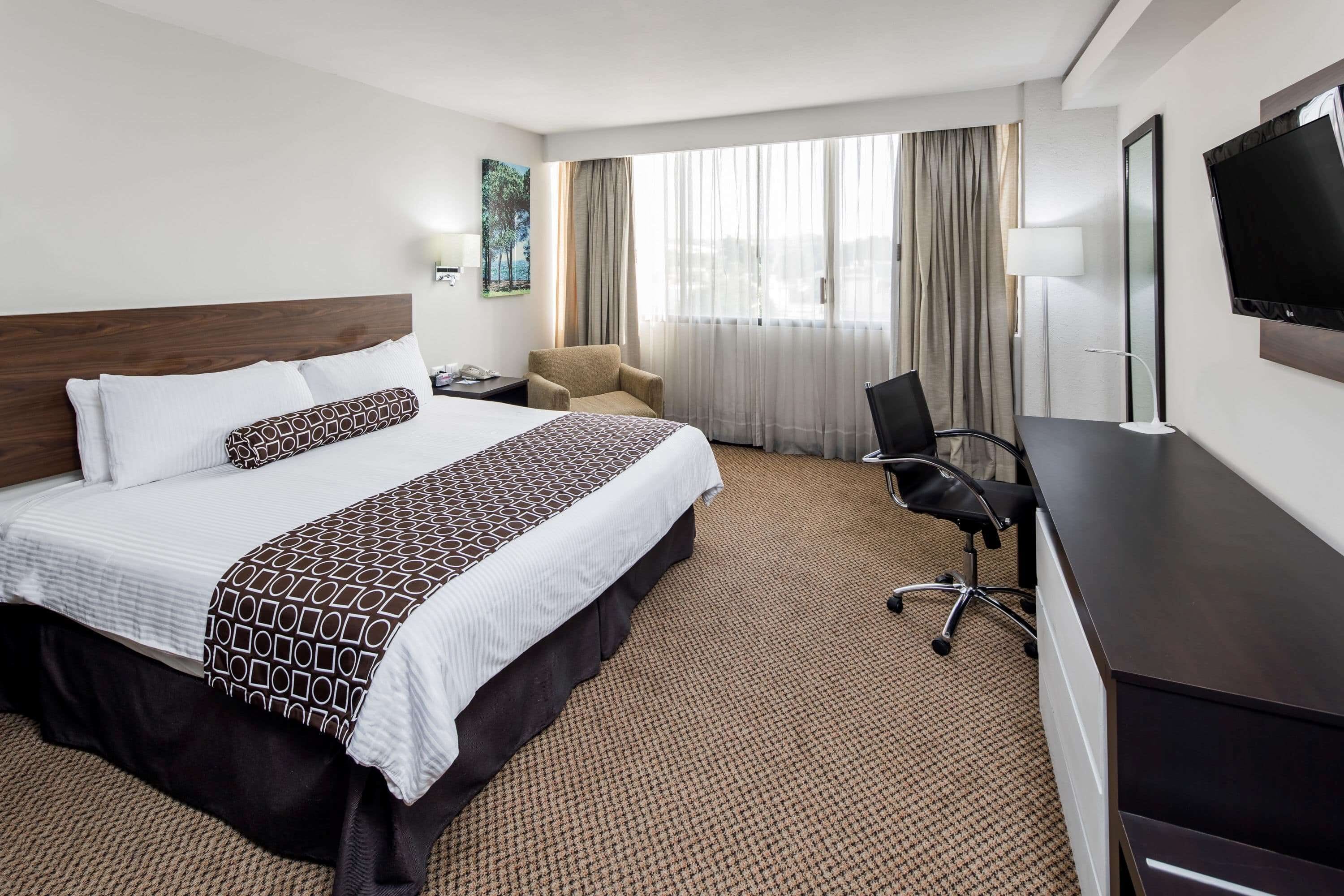 Une chambre d’hôtel avec un grand lit, un bureau, une chaise, une télévision et une fenêtre avec des rideaux.