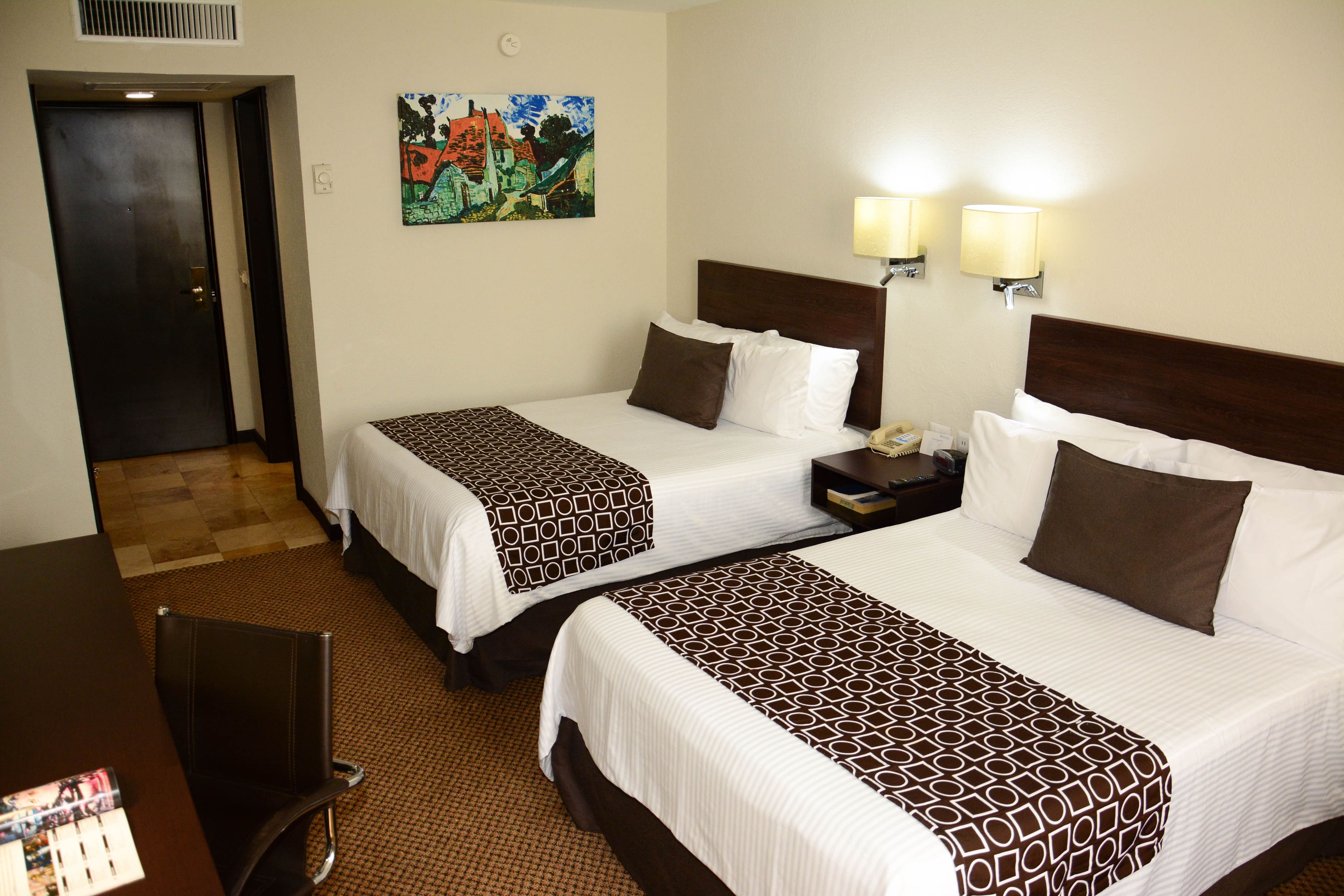 A hotel room with two beds, a desk, and a chair. There are two wall-mounted lamps and a painting on the wall.