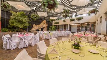 Un salón de banquetes con mesas redondas dispuestas para comer, sillas blancas y plantas colgantes.
