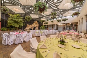 Un salón de banquetes con mesas redondas dispuestas para comer, sillas blancas y plantas colgantes.