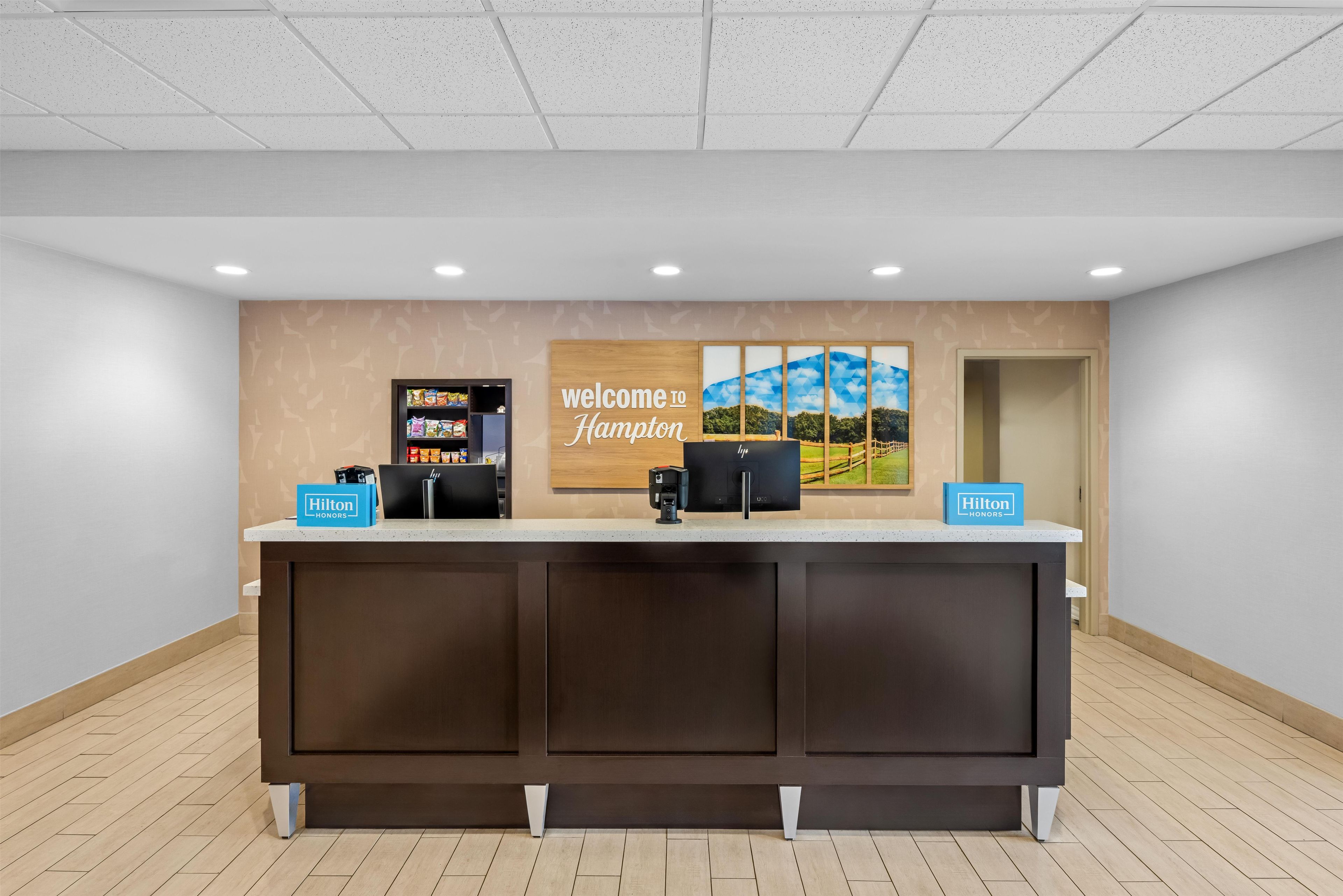 A reception desk with a 'Welcome to Hampton' sign, a minibar, and a 'Hilton Honors' sign.