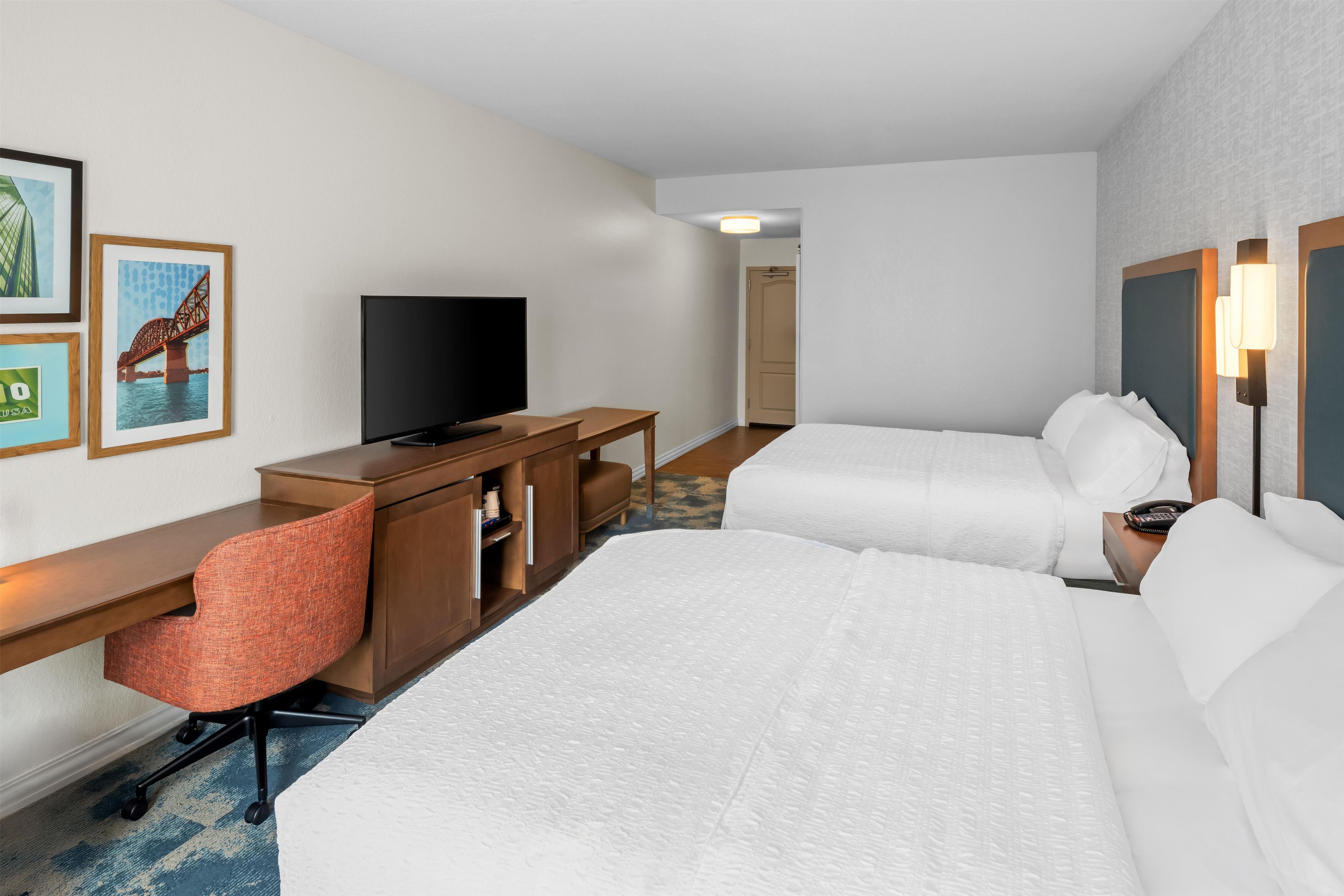 A hotel room with two beds, a desk with a television, a chair, and a door leading to another room.