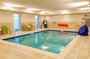 En indendørs swimmingpool med krystalklart vand, grønne stole, en badevagtstand og et håndklædestativ.