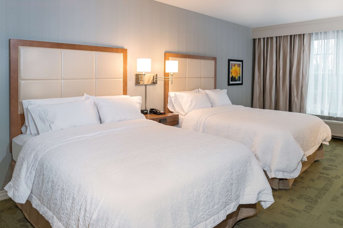 A hotel room with two beds, a nightstand with a lamp, a painting on the wall, and a window with curtains.