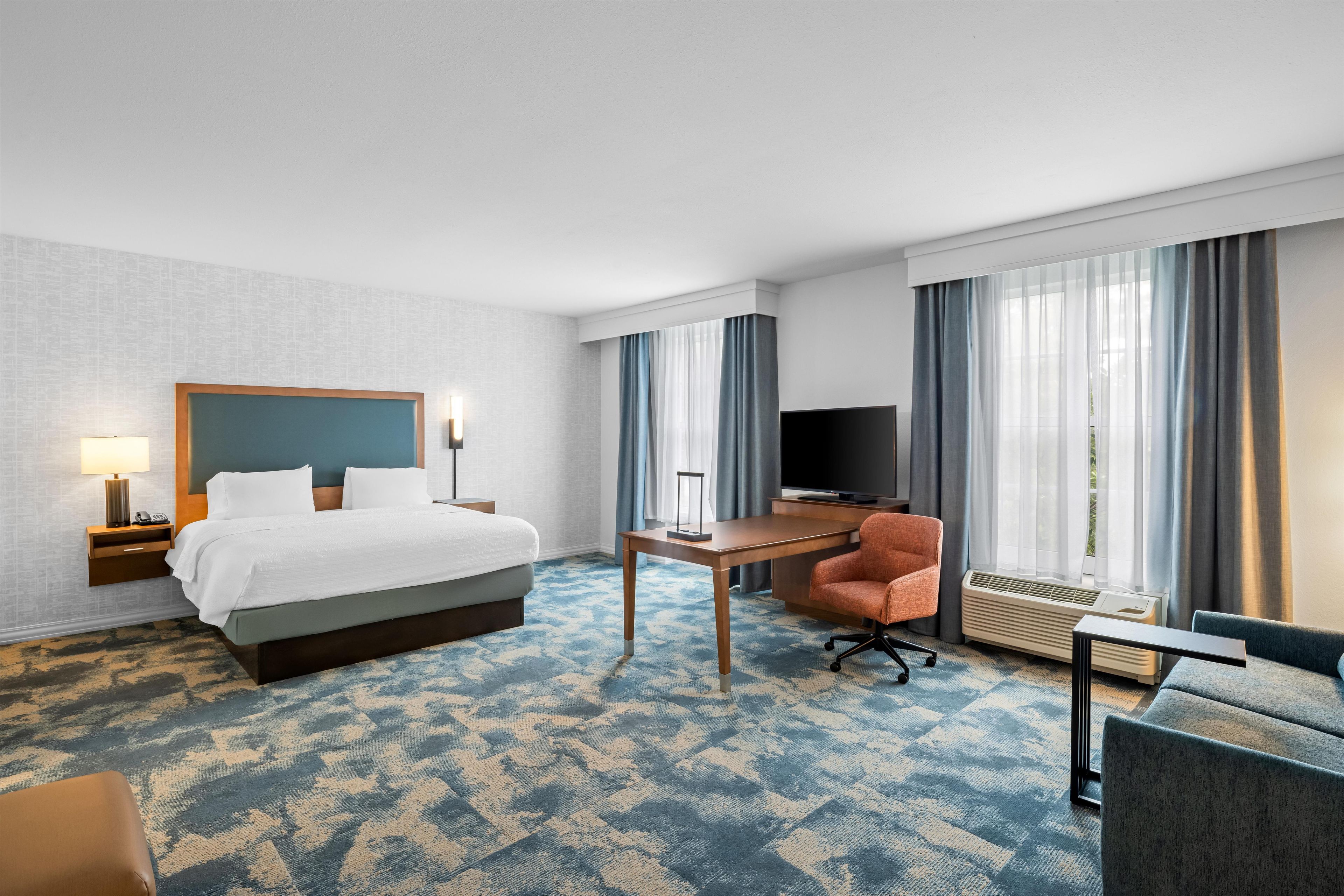 A hotel room with a bed, a desk, a chair, a TV, and a window with curtains.