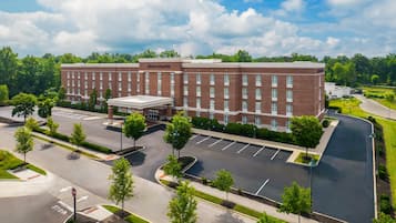 A large brick building with a central entrance and multiple windows, surrounded by a parking lot and greenery.