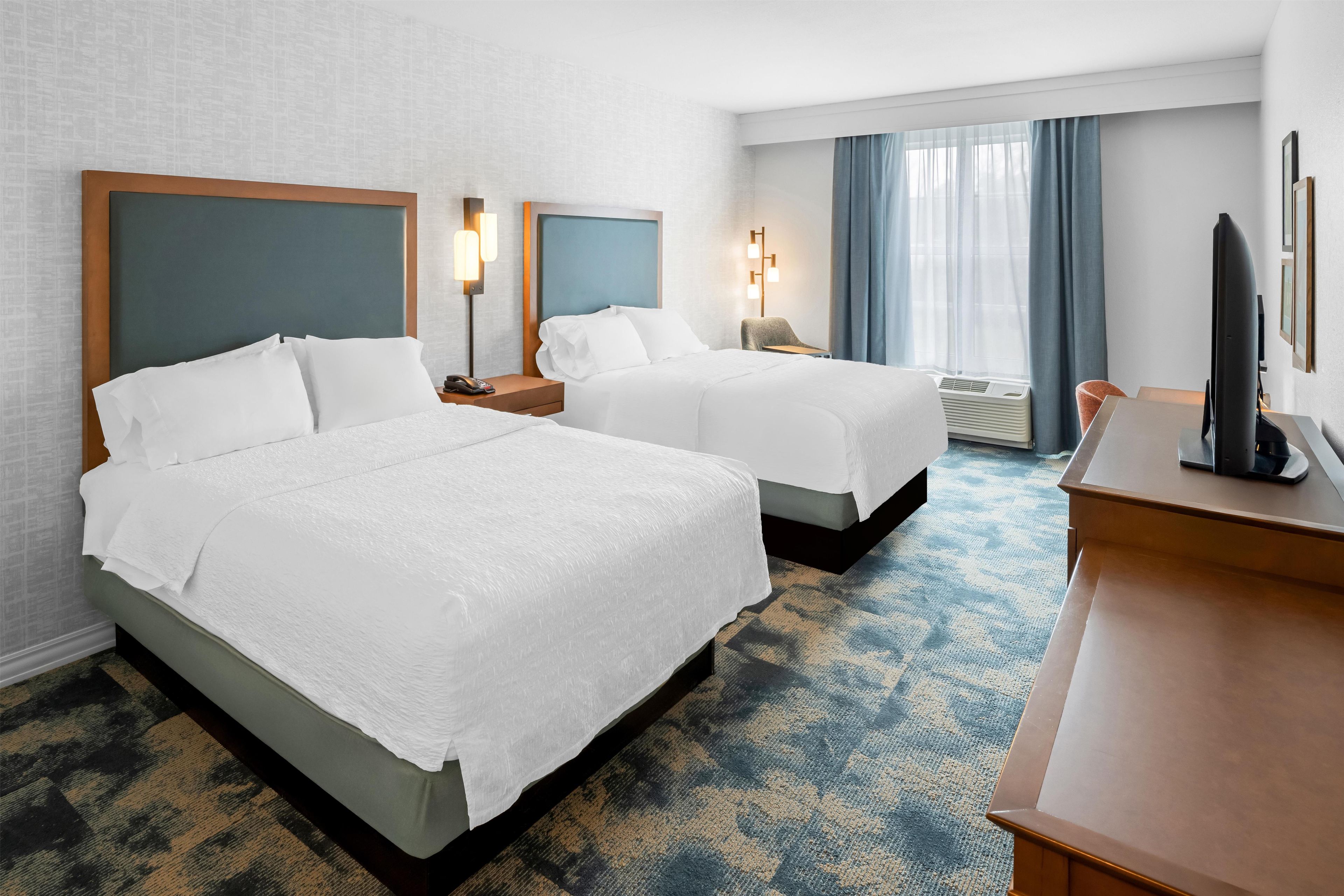 A hotel room with two beds, a desk, a chair, a lamp, and a large window with curtains.