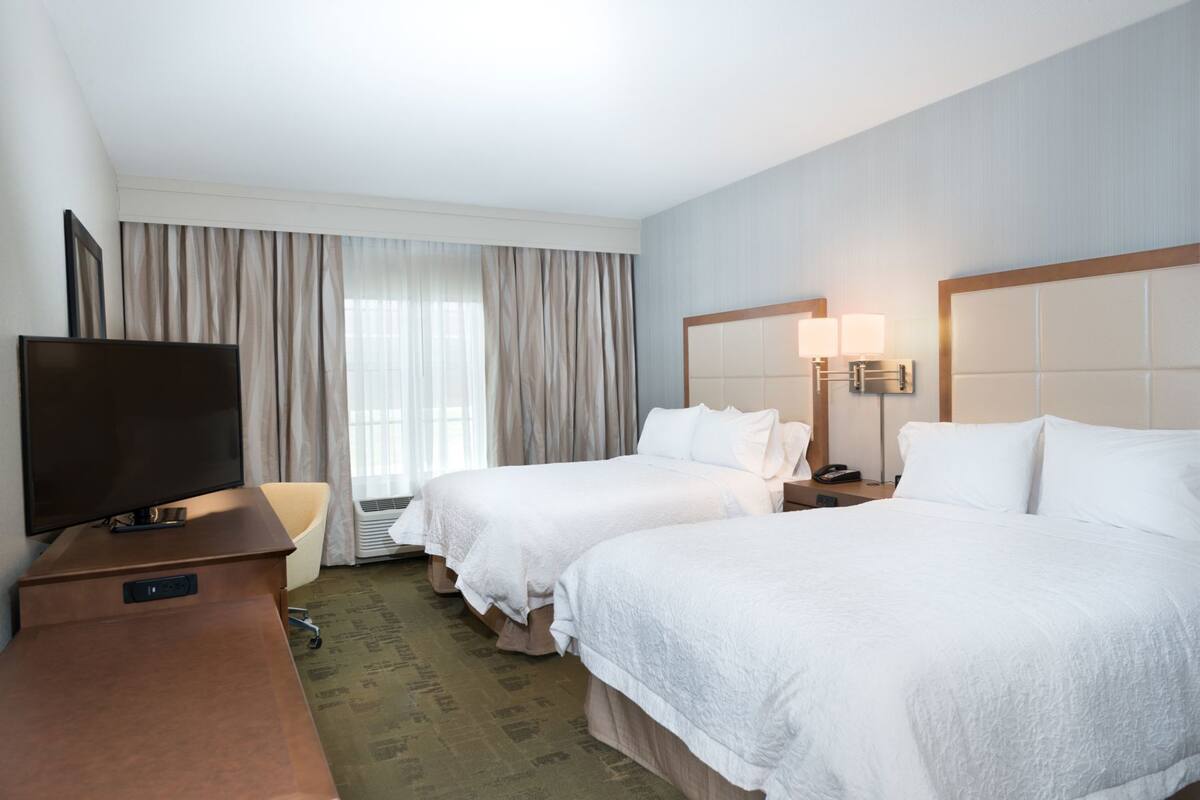 A hotel room with two beds, a TV, a desk, and a window with curtains.