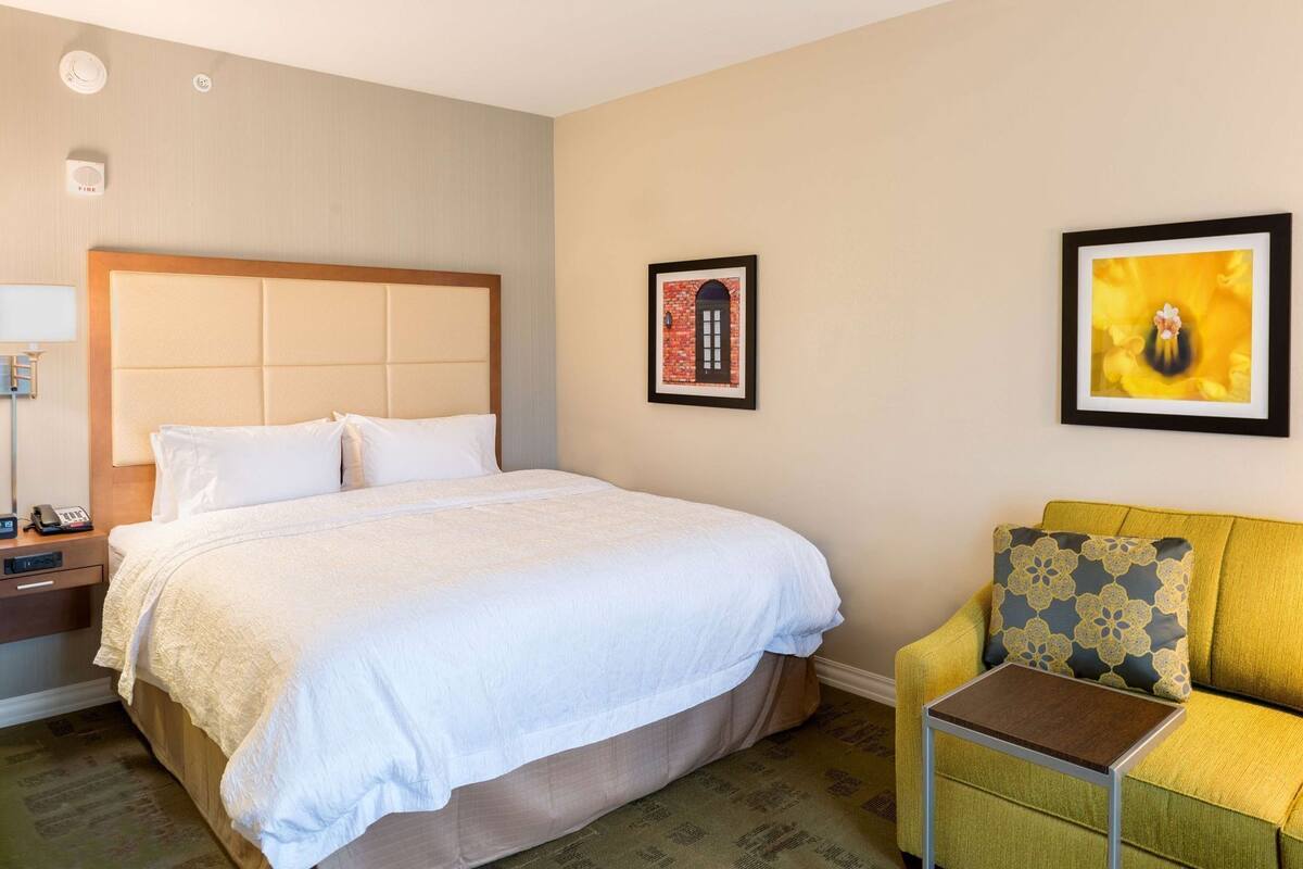 A hotel room with a bed, a nightstand, a chair, and two framed pictures on the wall.
