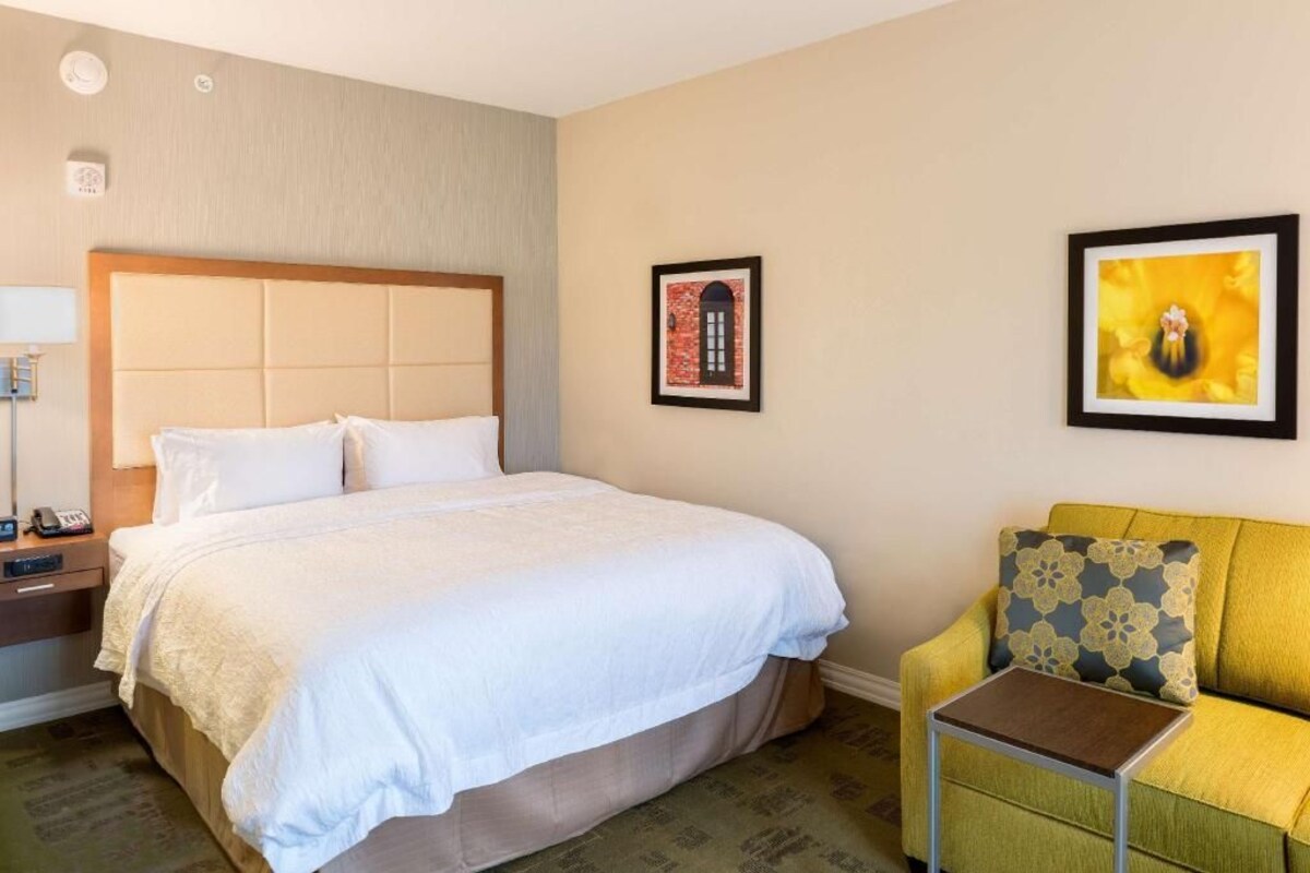 A hotel room with a bed, bedside table, lamp, and a sofa with a cushion.