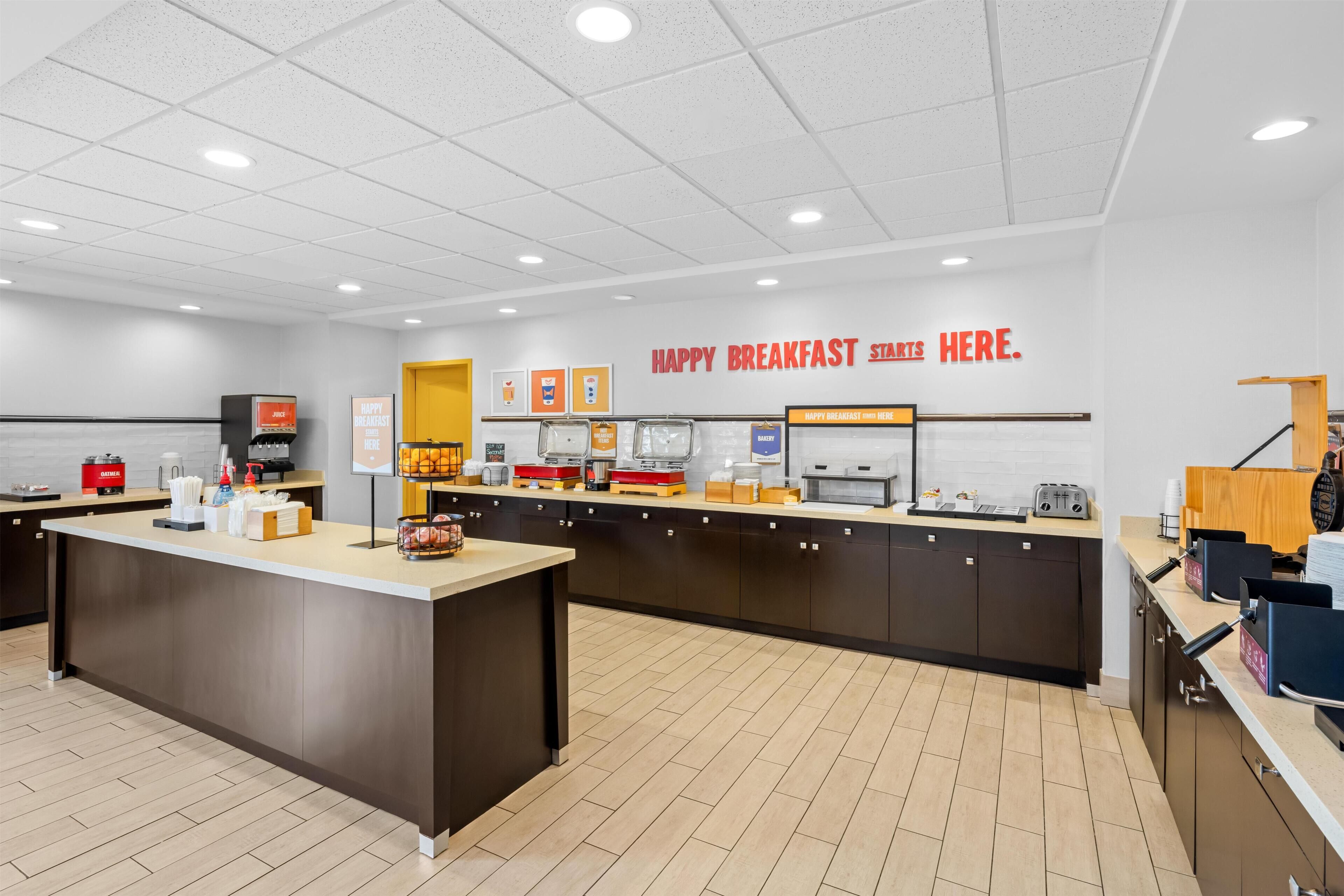 Un espace réservé au petit-déjeuner, avec un comptoir central, différents points de service où sont proposés divers aliments, et un panneau indiquant l’emplacement du service de petit-déjeuner.