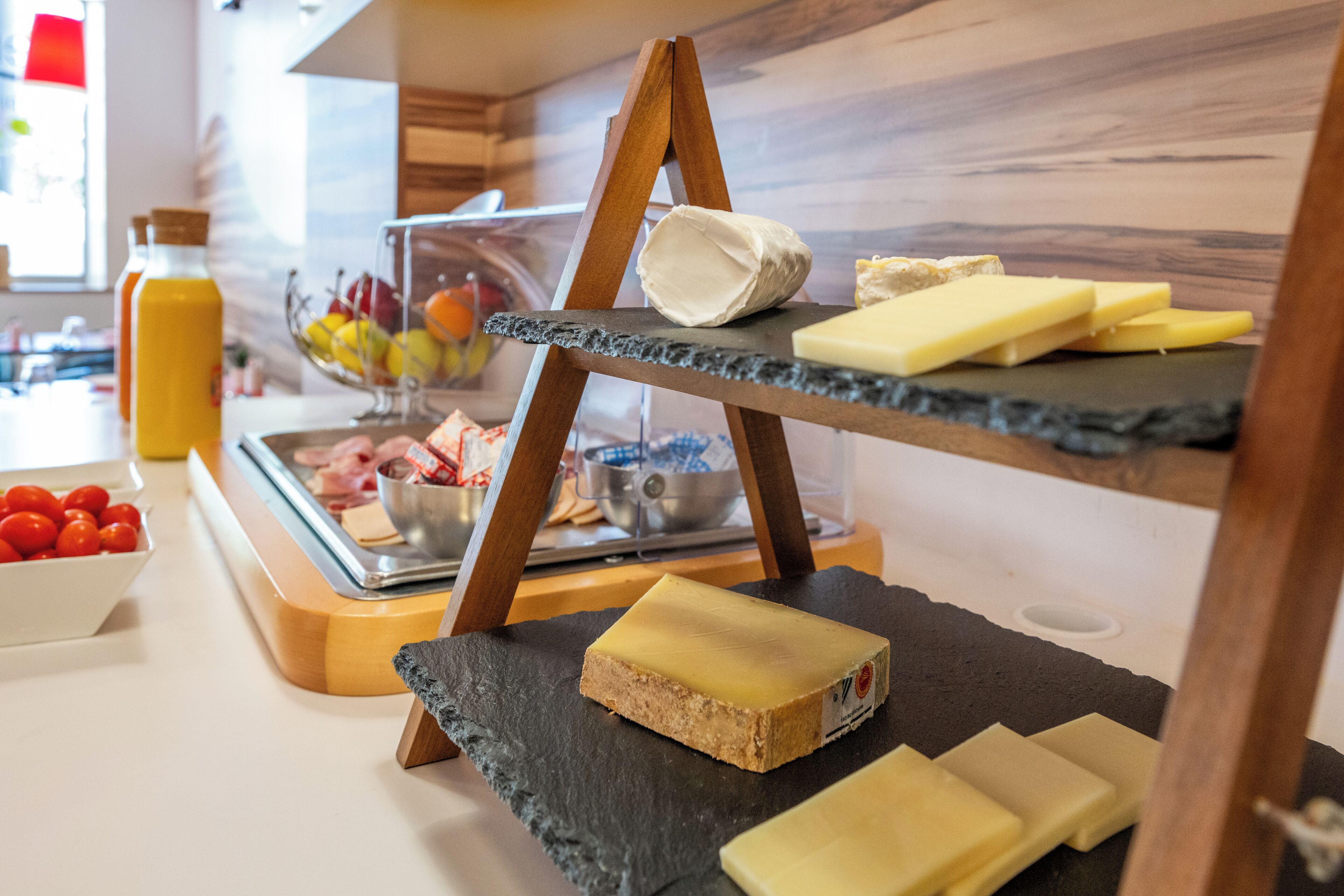 A cheese board with various cheeses and a bowl of fruits on a kitchen counter.