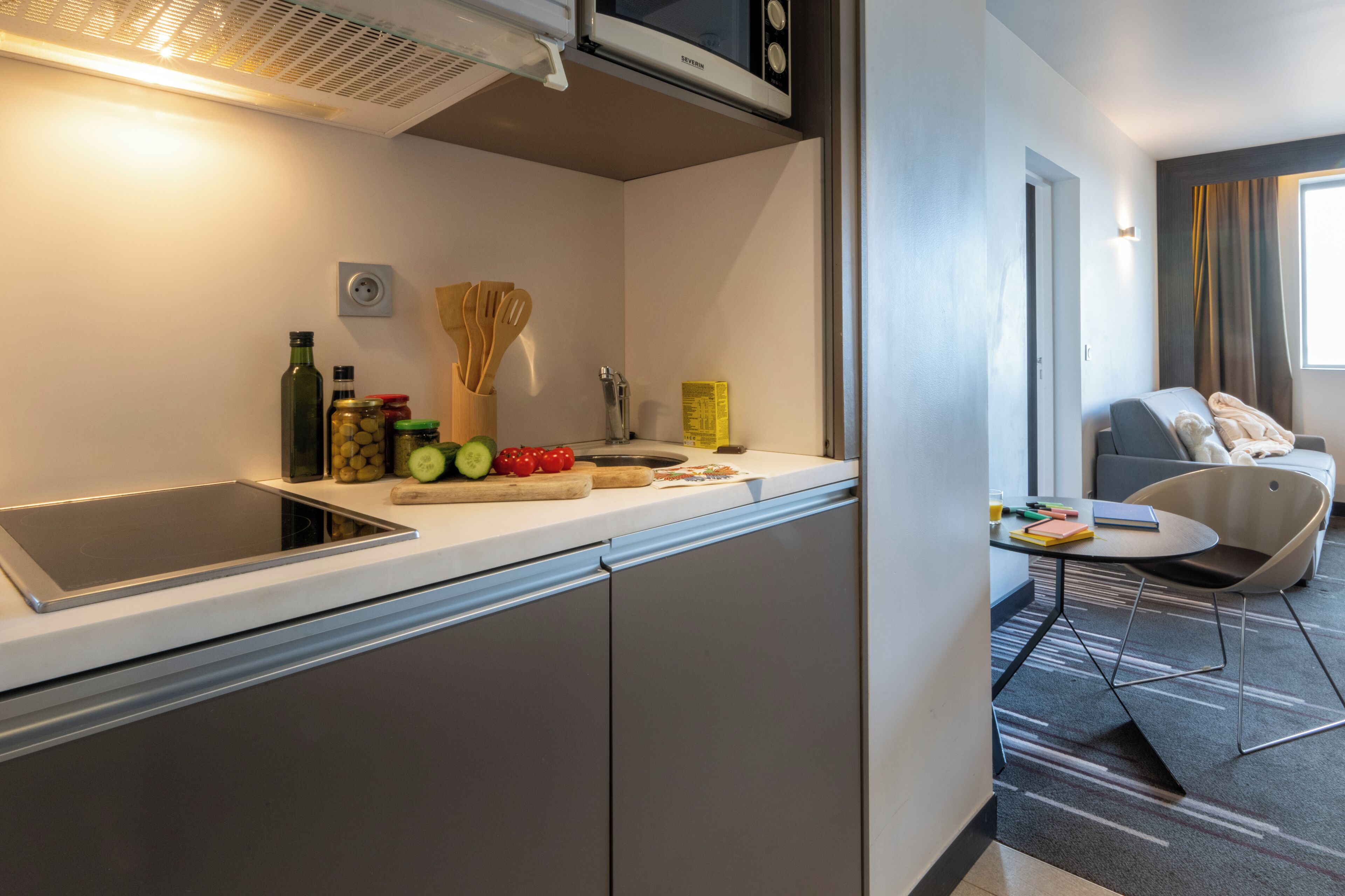 A modern kitchen with a built-in stove, a microwave, and a sink. There are fresh fruits and vegetables on the counter, and a small table with a chair.