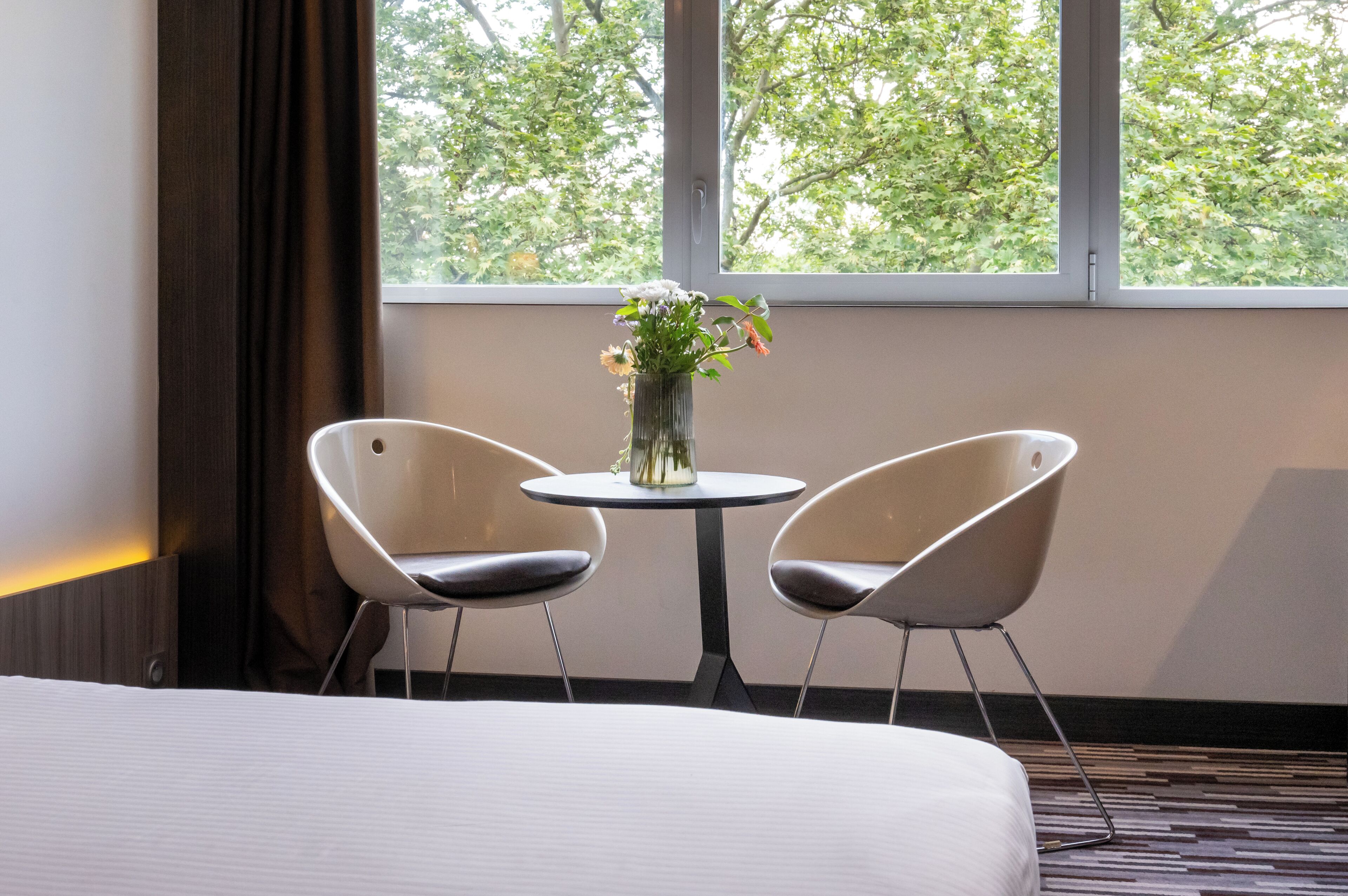 Habitación de hotel con una cama, dos sillas modernas, una mesita redonda con un jarrón de flores y una ventana con vistas a árboles.