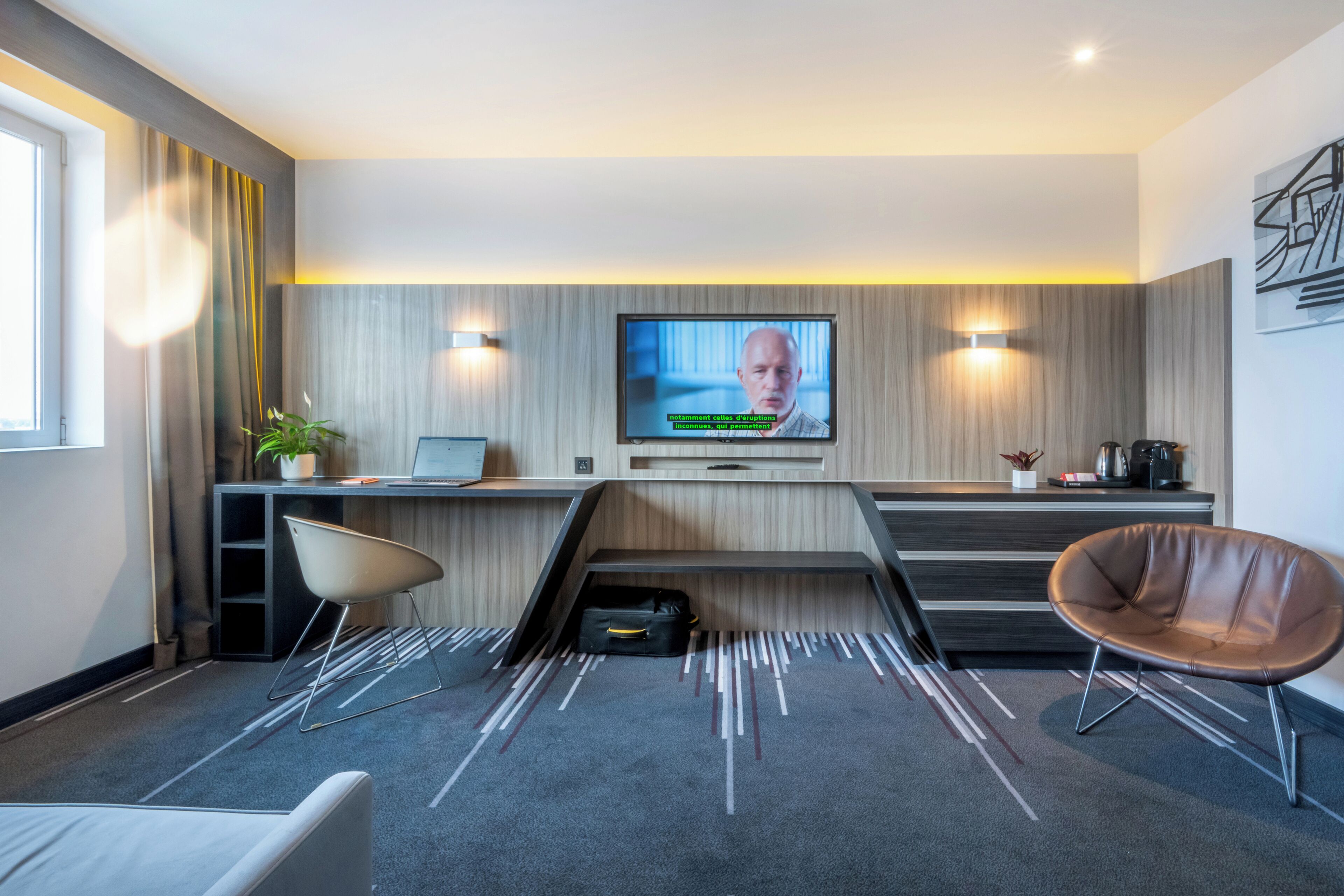 Ett modernt hotellrum med en platt-TV som är monterad på väggen, ett skrivbord med en bärbar dator, en stol och en soffa.