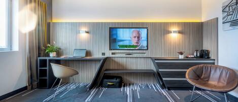 A modern hotel room with a flat-screen TV mounted on the wall, a desk with a laptop, a chair, and a sofa.