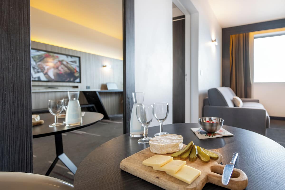 Una zona de comedor con una mesa de madera, queso, pepinillos y un cuchillo. Hay copas de vino y un bol sobre la mesa.