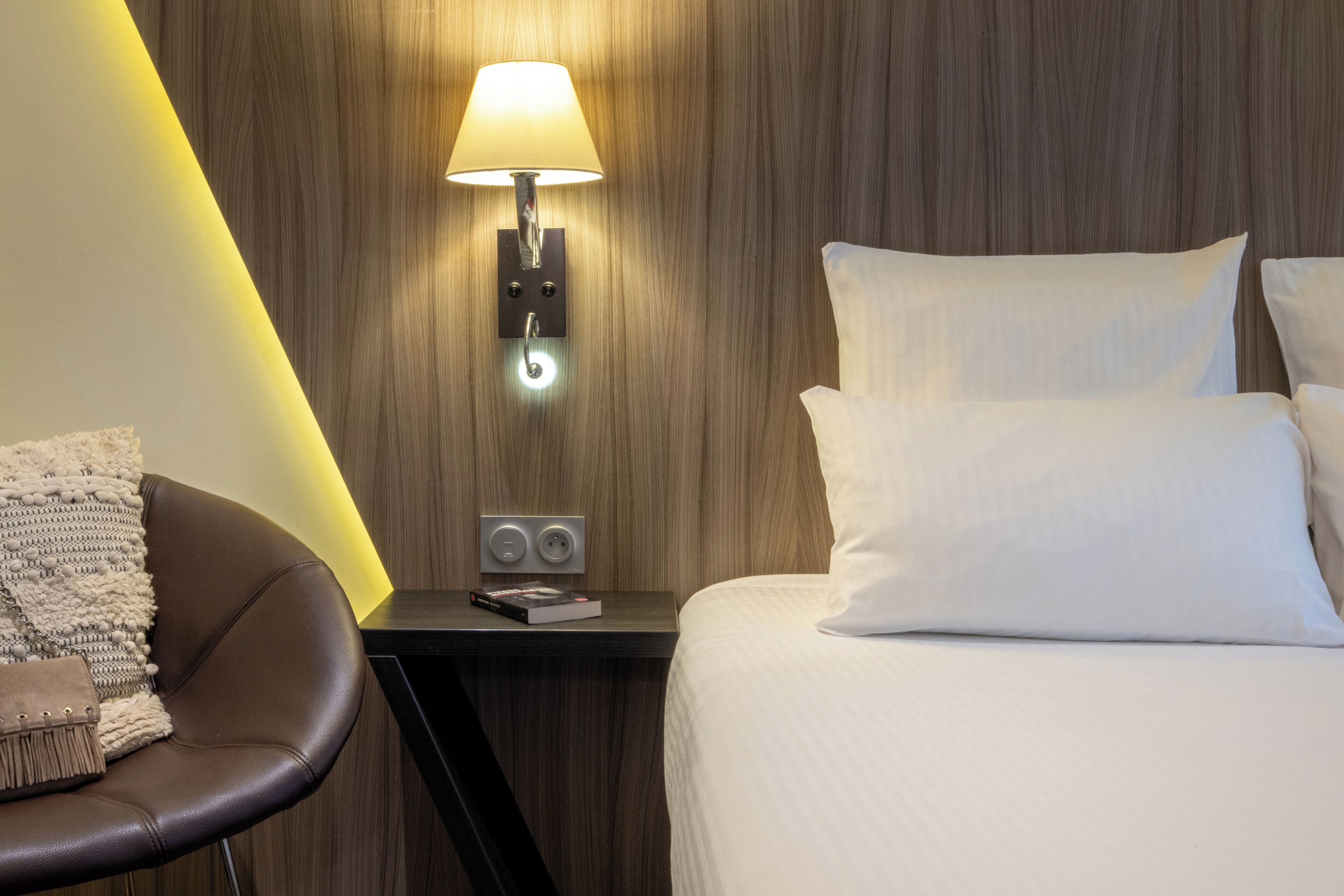 A hotel room with a bed, a nightstand with a lamp, a chair, and a wall-mounted control panel.