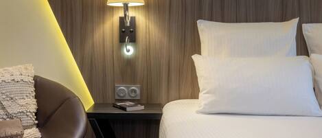 Habitación de hotel con cama, mesita de noche con lámpara, una silla y panel de control en la pared.