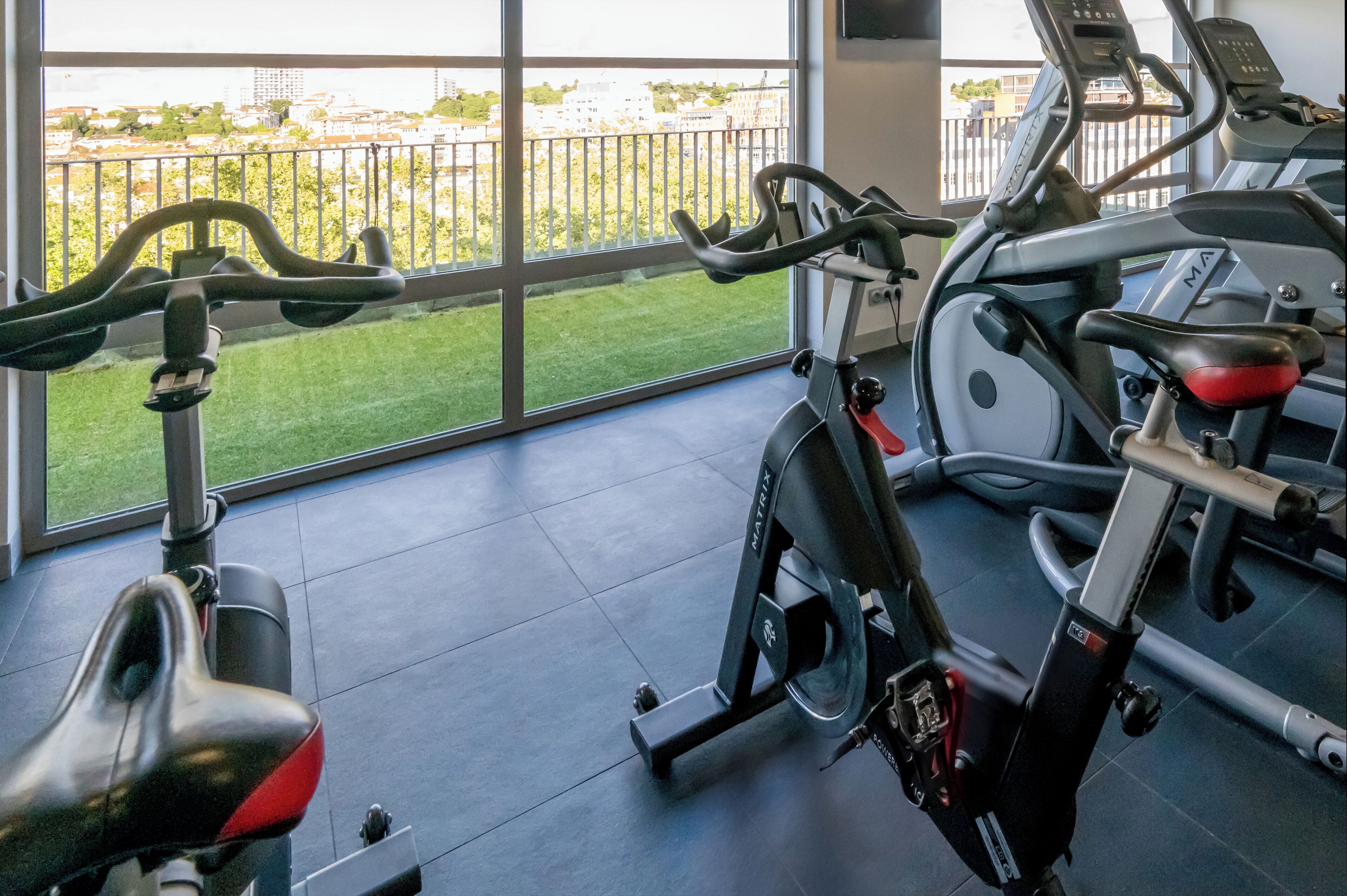 A gym with stationary bikes and a treadmill, large windows with a view of buildings and greenery.