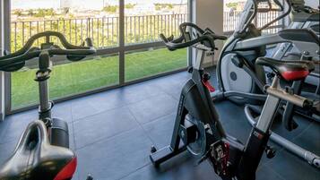 A gym with stationary bikes and a treadmill, large windows with a view of buildings and greenery.