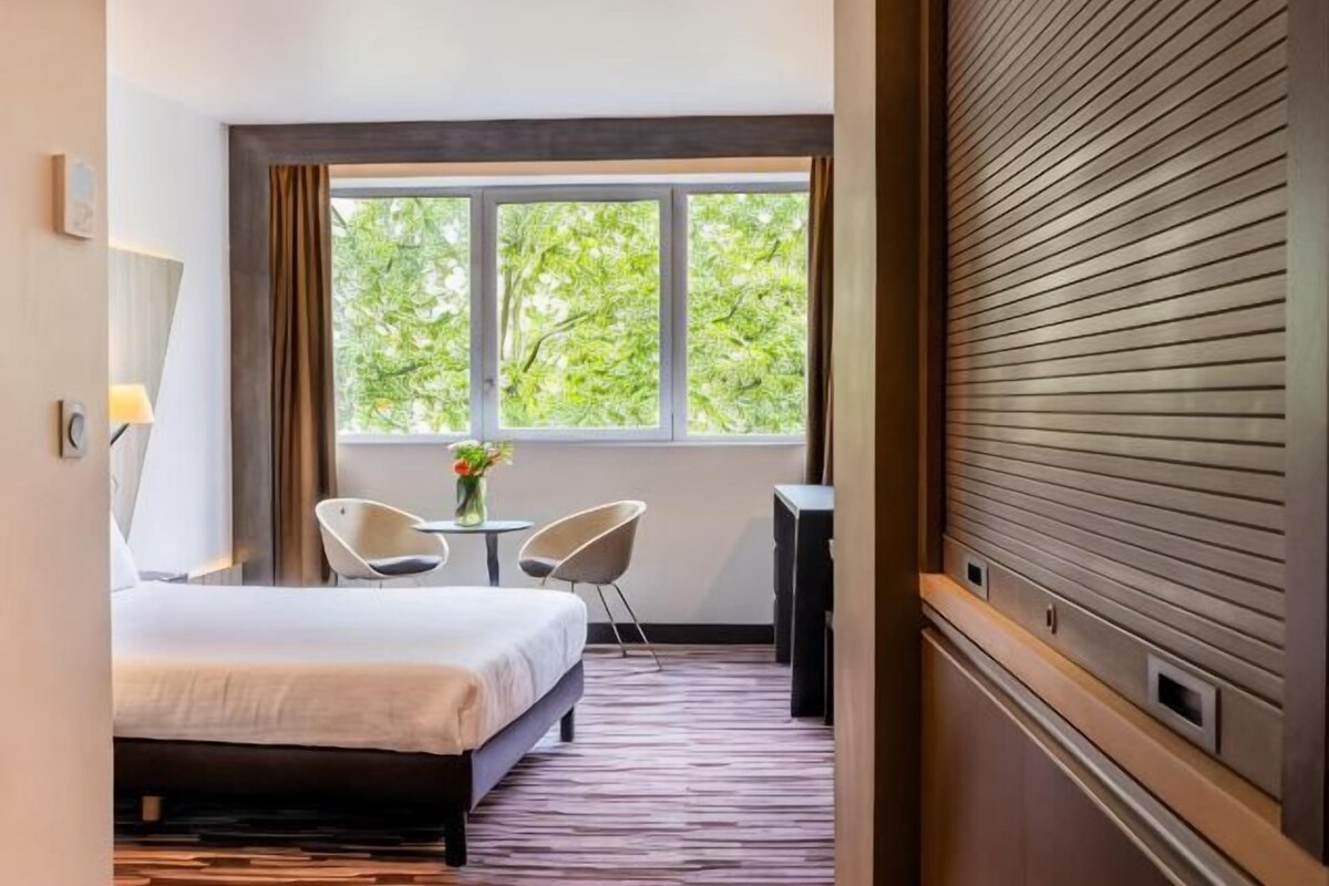 Una habitación de hotel con una cama, dos sillas, una mesita con un jarrón y una ventana con vistas a árboles.