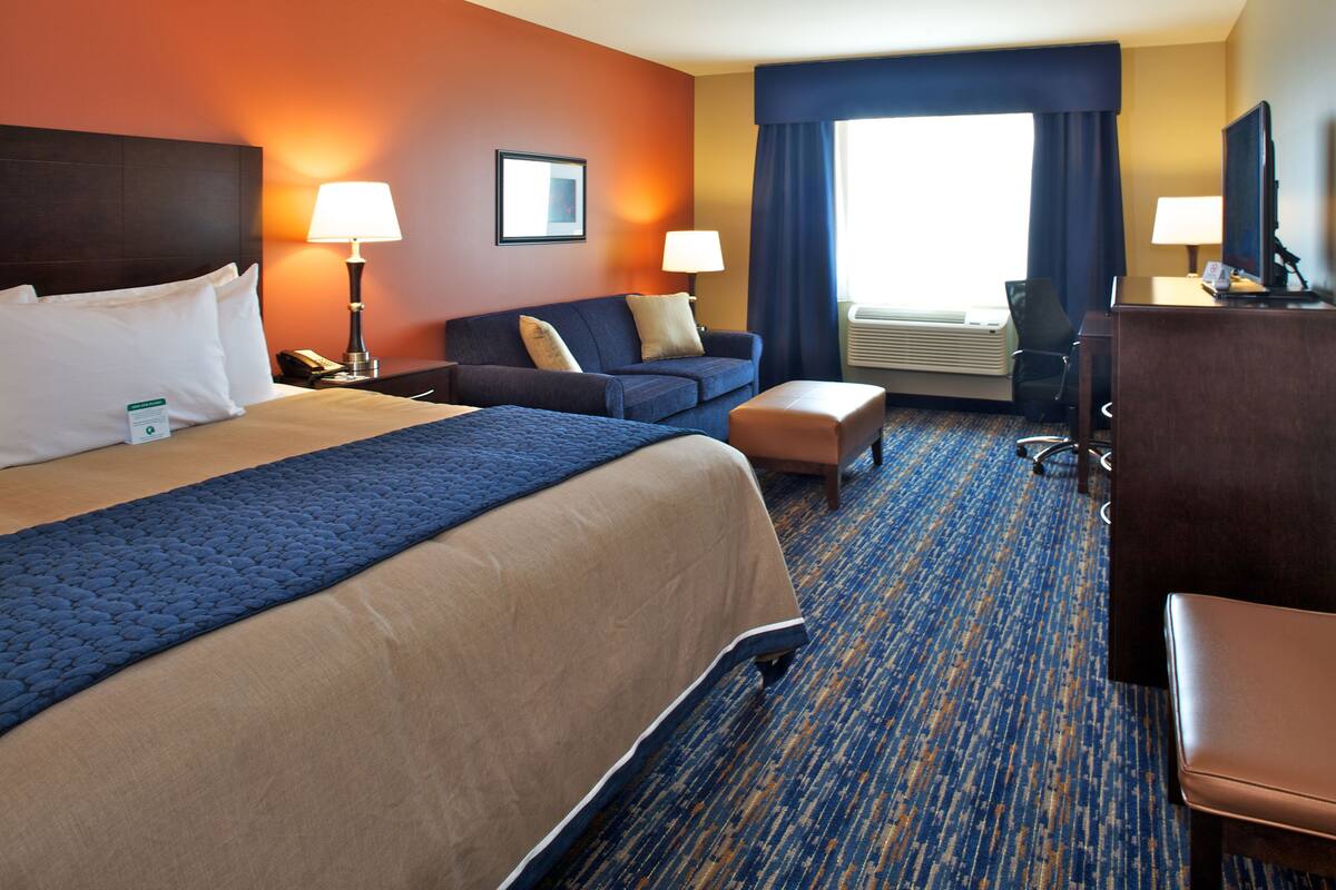 A hotel room with a large bed, a sofa, a desk, and a chair. There are lamps on the nightstands and a large window with curtains.