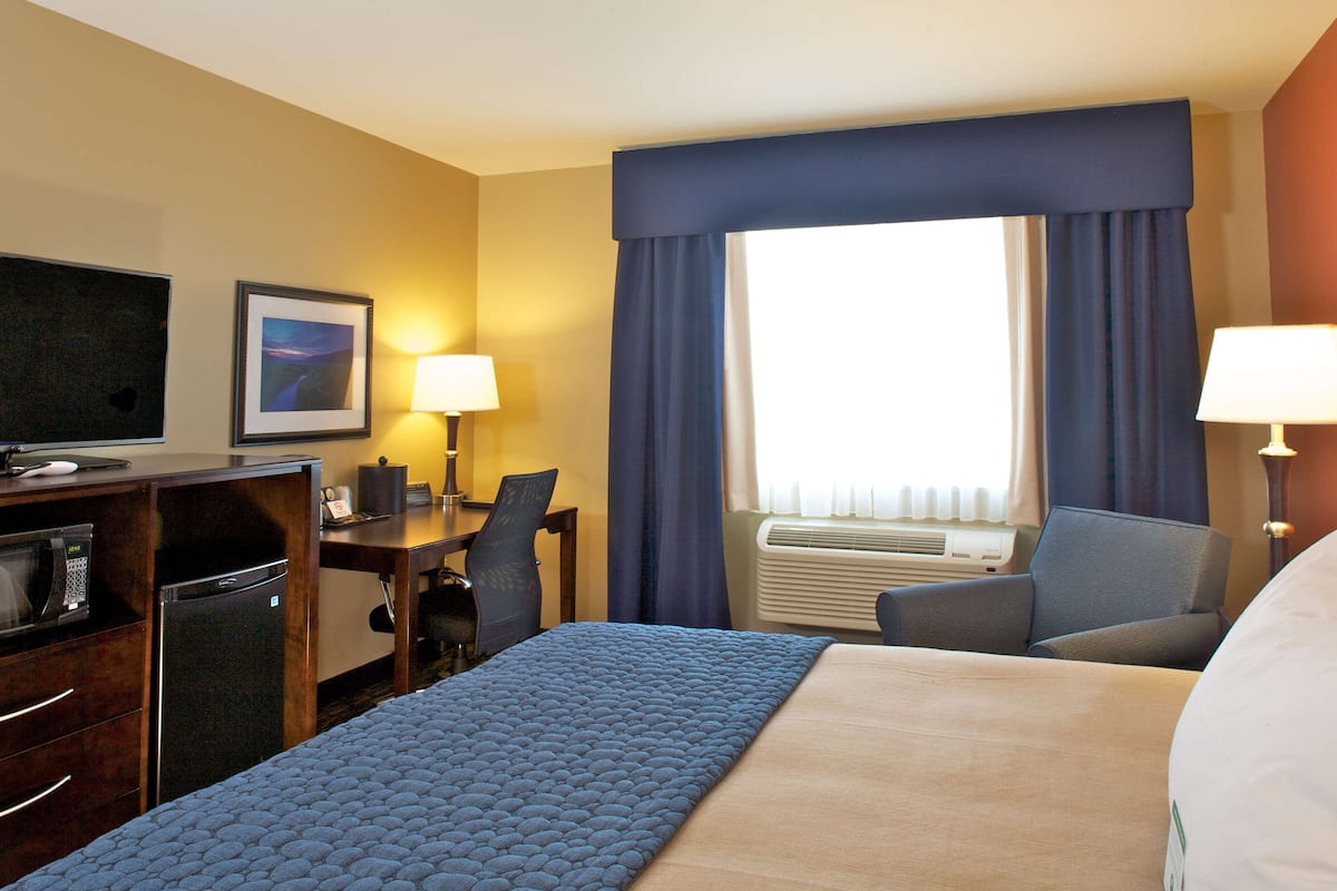 A hotel room with a bed, desk, chair, television, air conditioning unit, and a window with curtains.