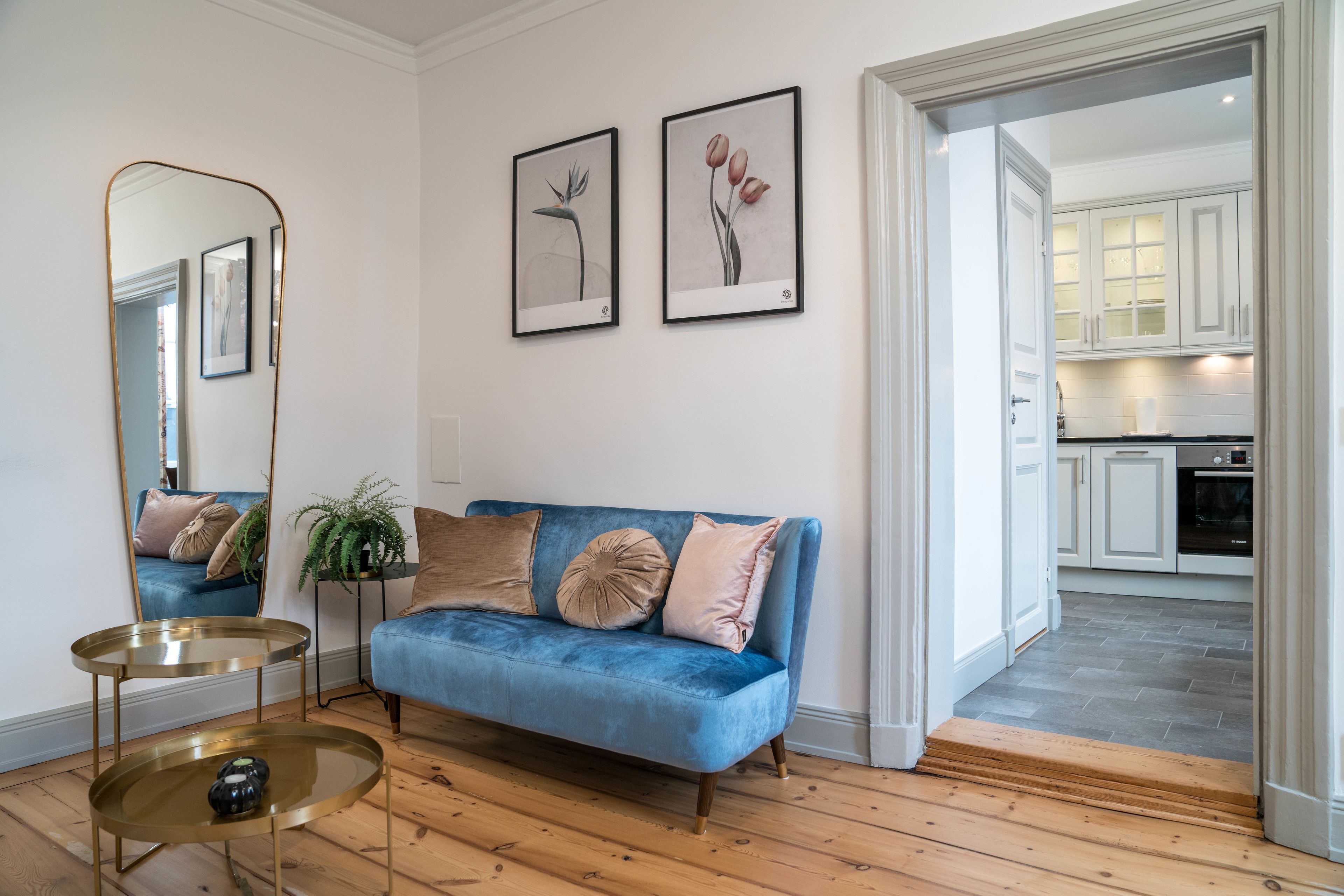 A living room with a blue sofa, a round coffee table, and a mirror. There are two framed pictures on the wall, and a plant on a side table.