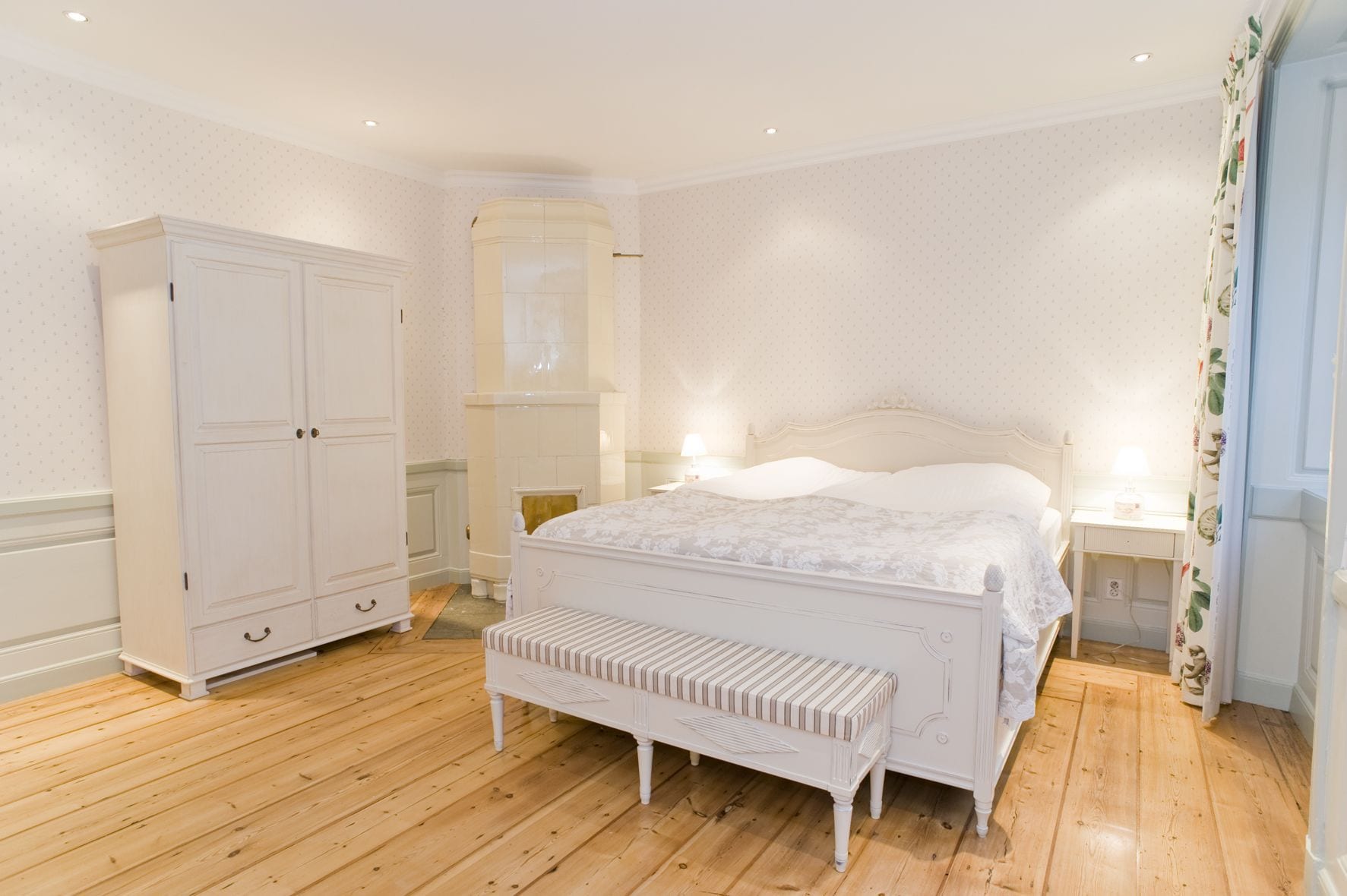 A bedroom with a large bed, a wardrobe, a bench, and a nightstand with a lamp.