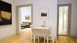 Una habitación con piso de madera, una mesa blanca con sillas, un cuadro en la pared y un umbral que conduce a otra habitación.
