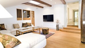 Una sala de estar moderna con un sofá blanco, vigas de madera, un televisor de pantalla plana y una escalera de madera que conduce a otra habitación.