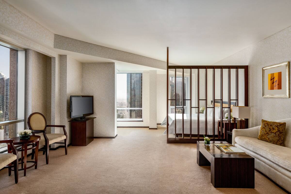 A modern hotel room with a large window, a flat-screen TV, a sofa, a coffee table, and a dining area with chairs and a small table.