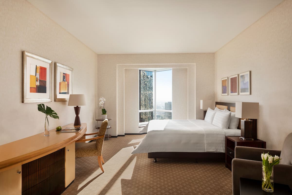 A hotel room with a large bed, a desk, a chair, a nightstand, and a window with a view of the city.