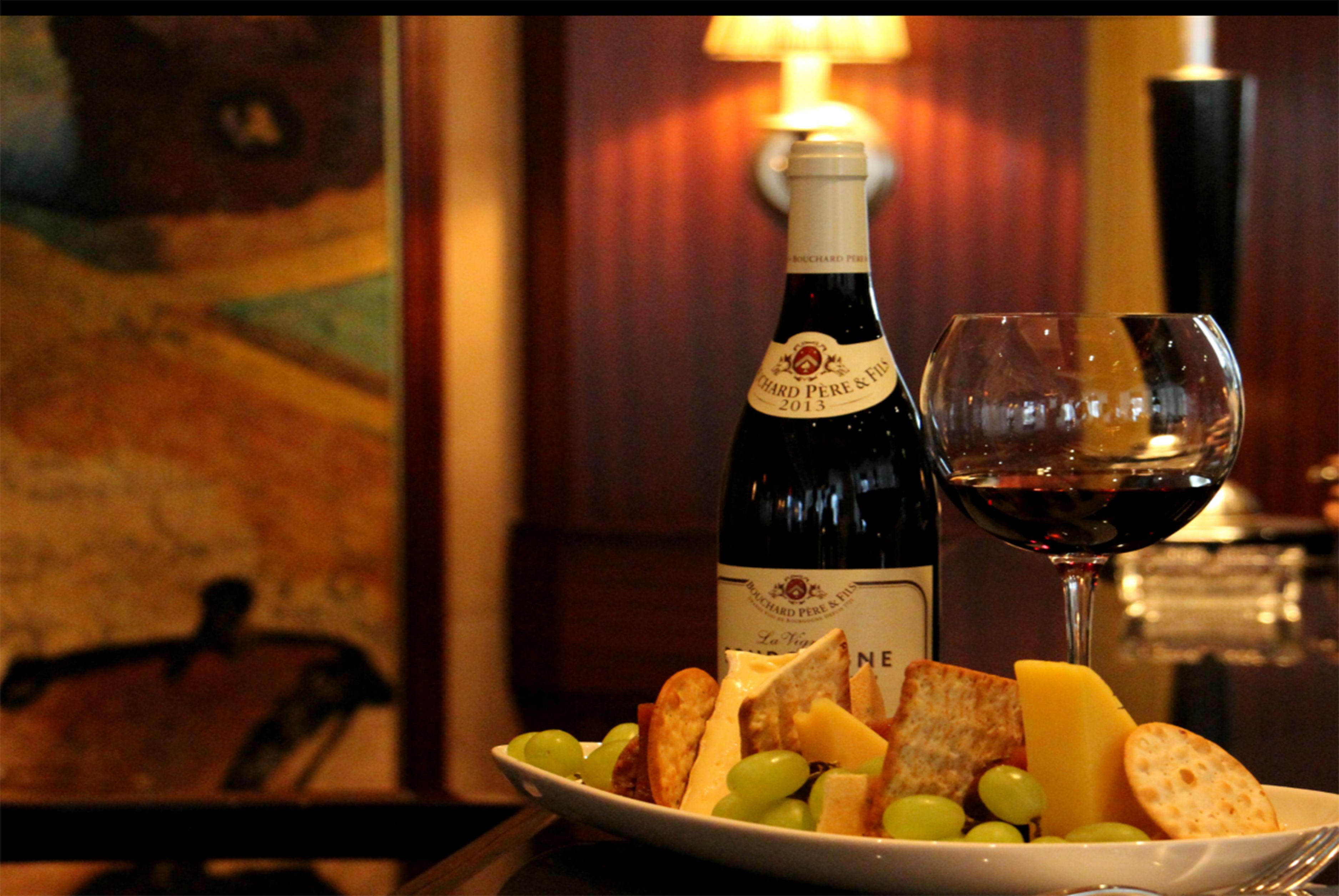 A bottle of wine, a glass of red wine, and a plate with cheese and grapes.
