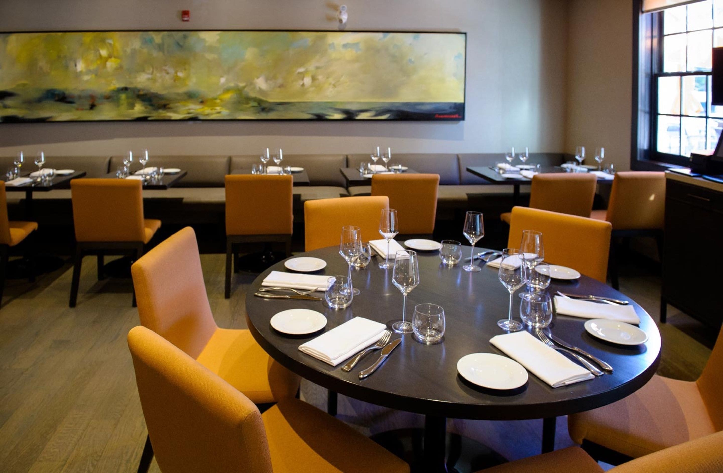 Comedor con mesas redondas dispuestas para cuatro comensales, cada una con platos blancos, cubiertos y copas de vino. Las sillas están tapizadas con un tejido naranja claro. Un cuadro abstracto de gran tamaño adorna la pared sobre las mesas.