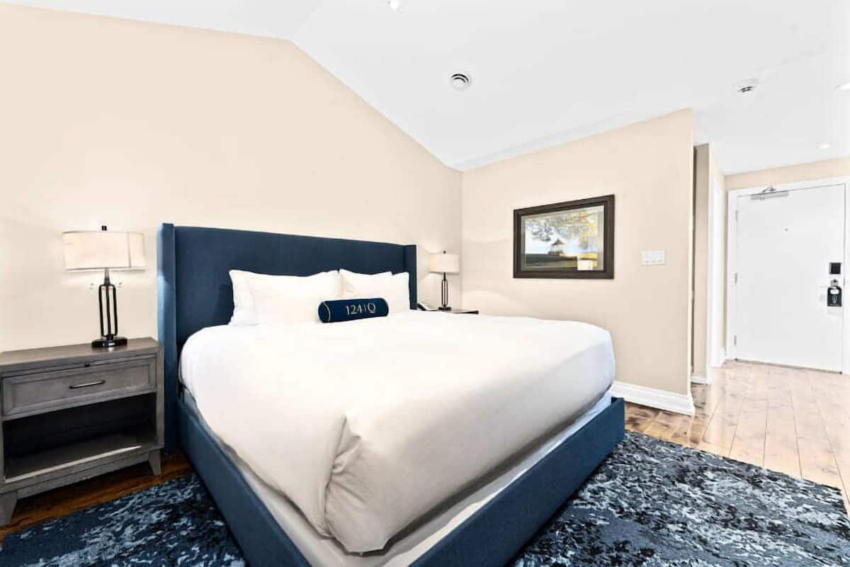 Une chambre d’hôtel avec un grand lit, une table de chevet, une lampe, un tableau encadré au mur et une porte.