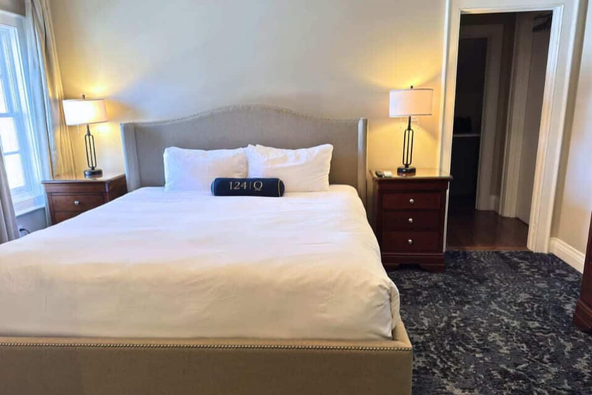 Une chambre d’hôtel avec un lit, des tables de chevet, une lampe, une horloge et un sol recouvert de moquette.