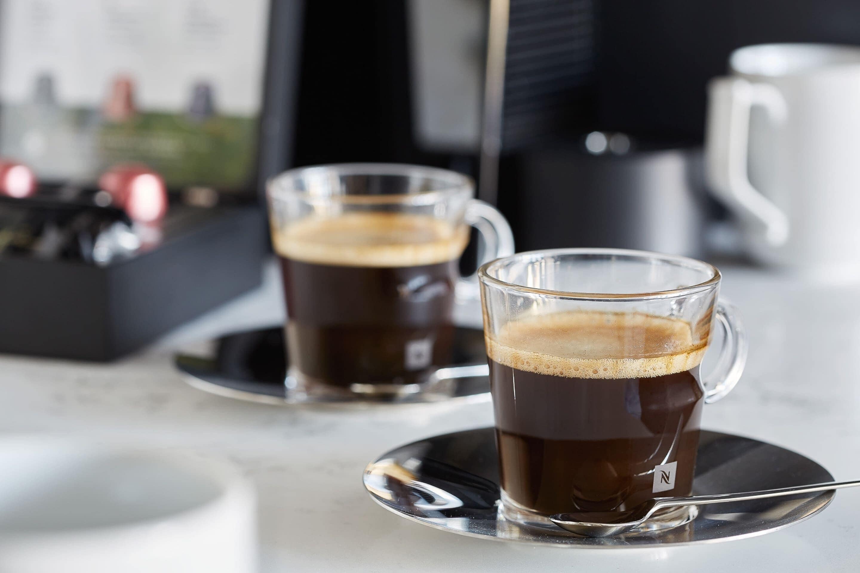 Twee heldere koffiekopjes met schoteltjes, een lepel, een witte mok en een koffieapparaat op de achtergrond.