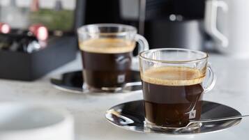 Twee heldere koffiekopjes met schoteltjes, een lepel, een witte mok en een koffieapparaat op de achtergrond.