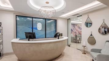 A reception desk with a modern design, a chandelier, a large window with a view, and a seating area with a sofa.
