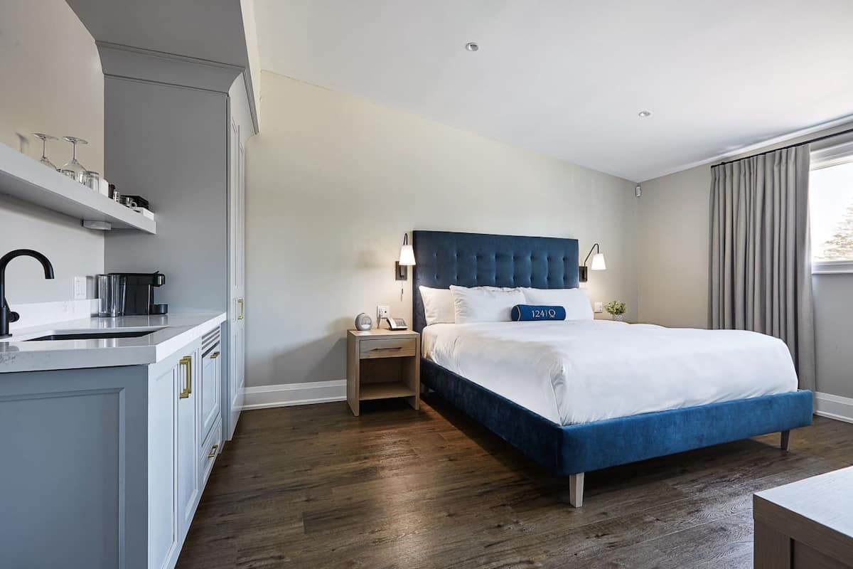 Une chambre d’hôtel moderne dotée d’un grand lit, d’une table de chevet, d’une commode, d’un coin cuisine intégré et d’une fenêtre avec des rideaux.