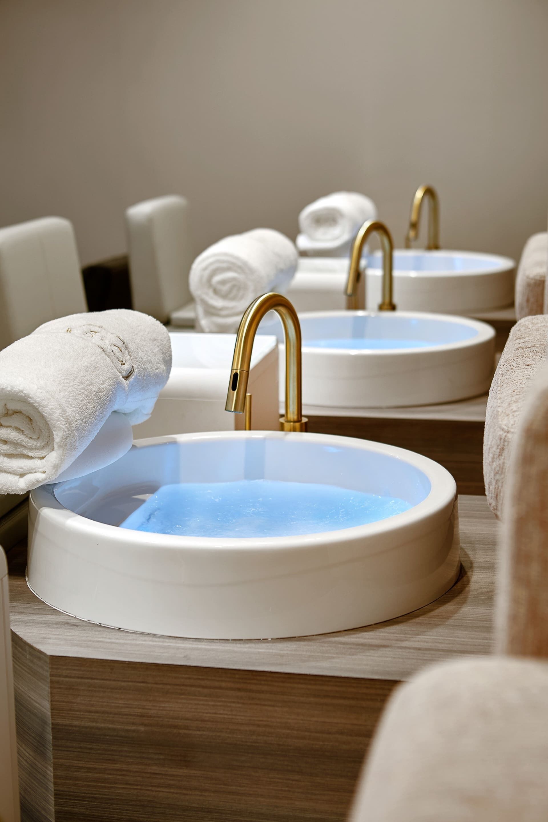 A bathroom with three sinks, each with a gold faucet and a white basin. Towel racks are visible in the background.