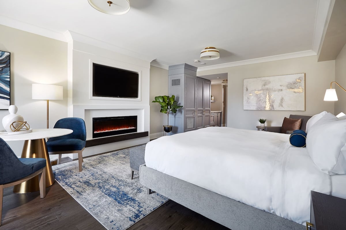 Une chambre d’hôtel moderne dotée d’un grand lit, d’un téléviseur à écran plat fixé au-dessus d’une cheminée, d’un coin salon avec une table ronde et des chaises, et d’une grande fenêtre.