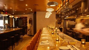 Una larga mesa de comedor con sillas, lámparas colgantes en el techo y una zona de cocina al fondo.